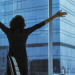 Back View of the Businesswoman Standing in Her Office Stretching, Raising Arms, Celebrating Success while Looking out of Window with Cityscape View.